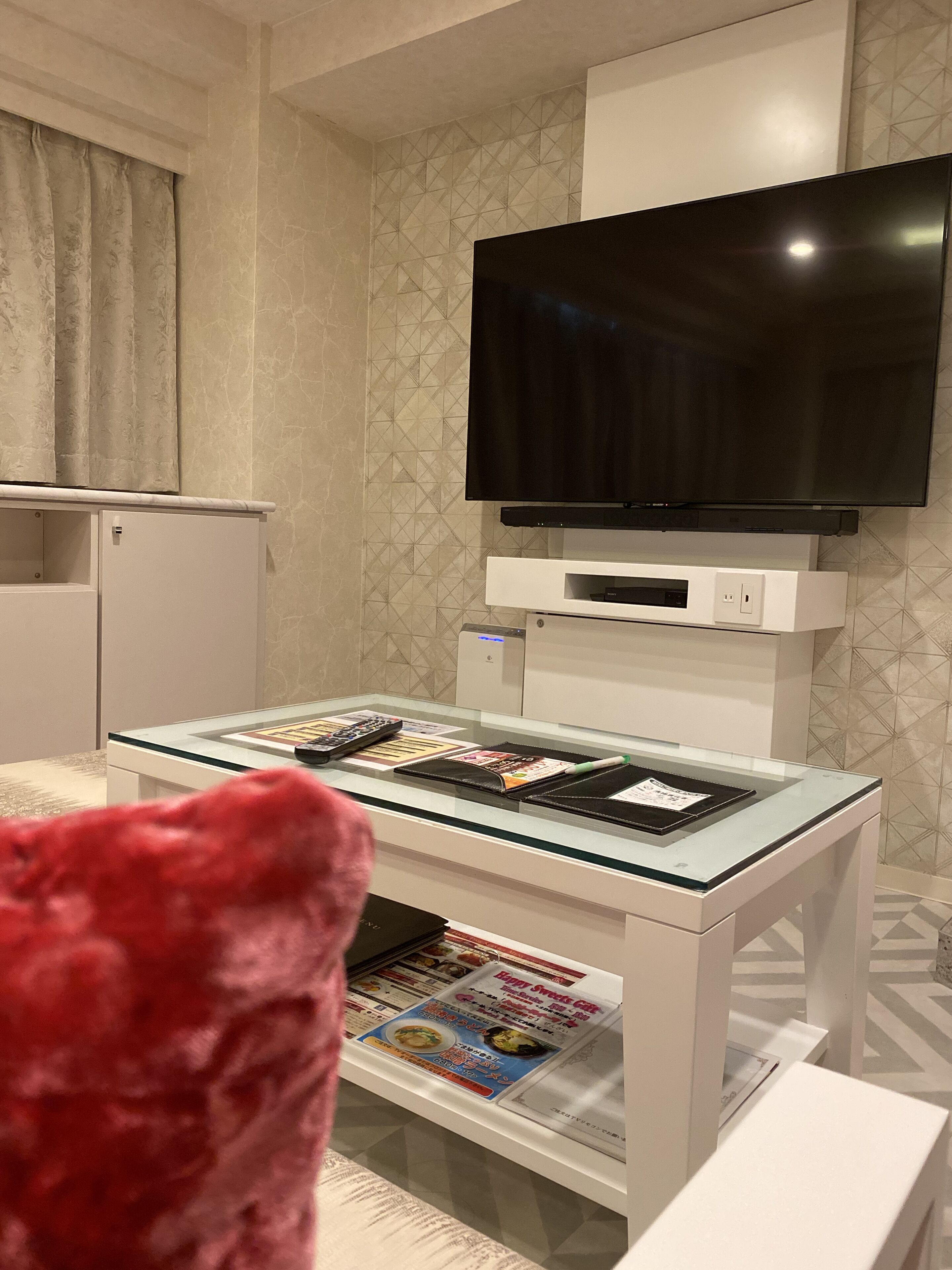 Een moderne woonkamer met een flatscreen televisie aan de muur, een salontafel met glazen blad en tijdschriften, een kleine koelkast en een rode fauteuil.