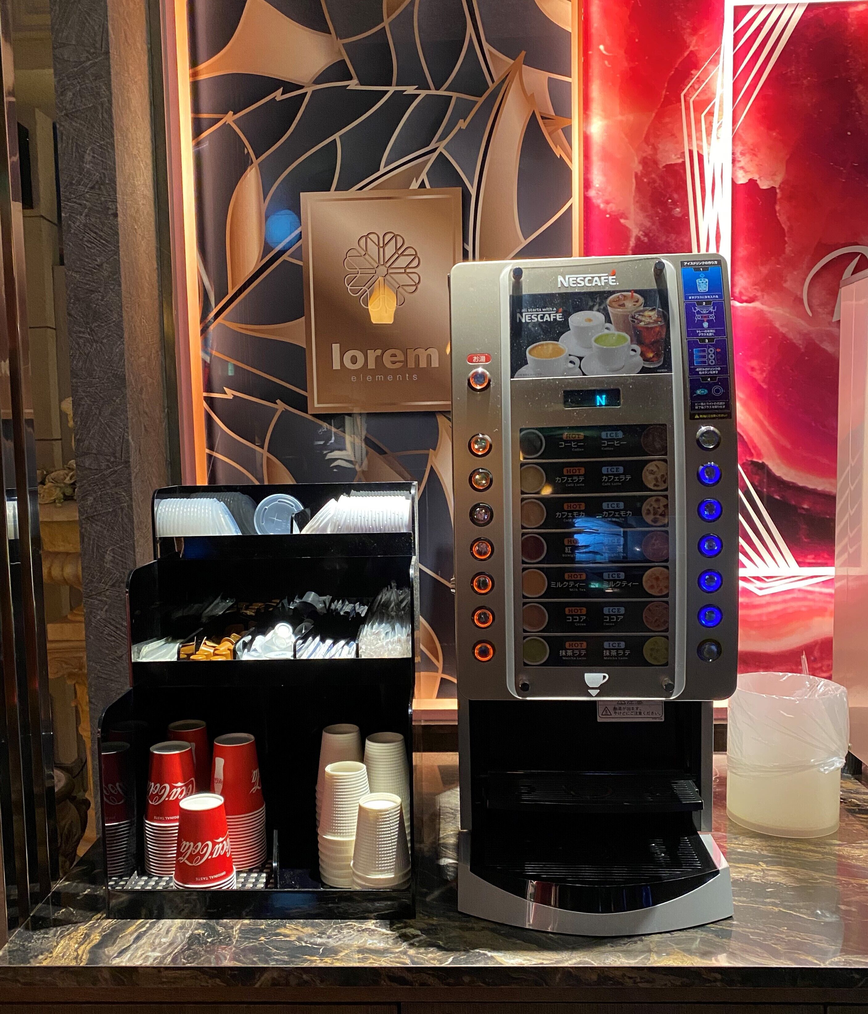 A coffee machine with a digital display and multiple buttons, a stack of disposable cups, and a Coca-Cola refrigerator.