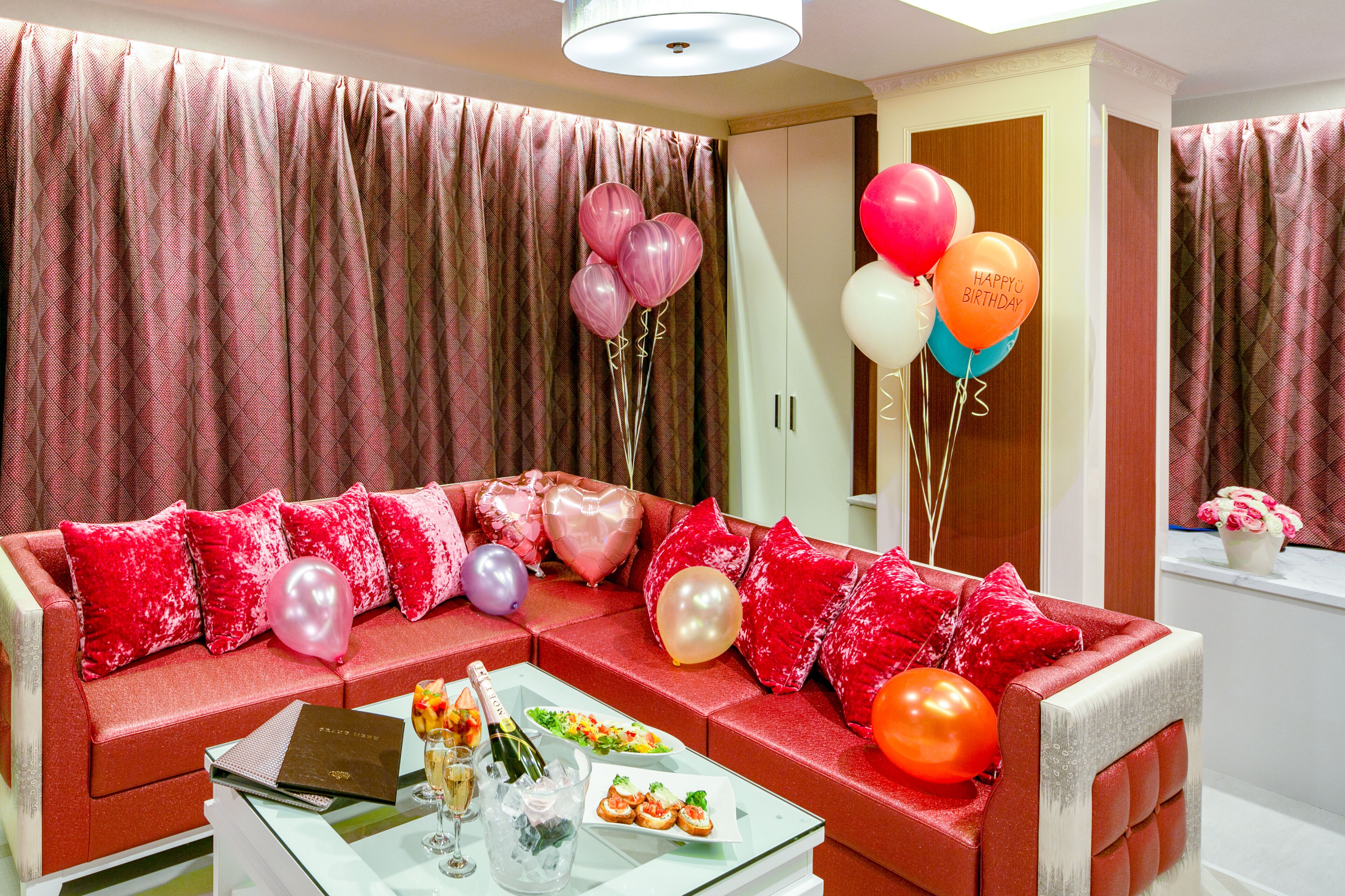 Un sofá rojo con globos rosa y morados, una mesa de cristal con una botella y aperitivos, y una puerta con un espejo.