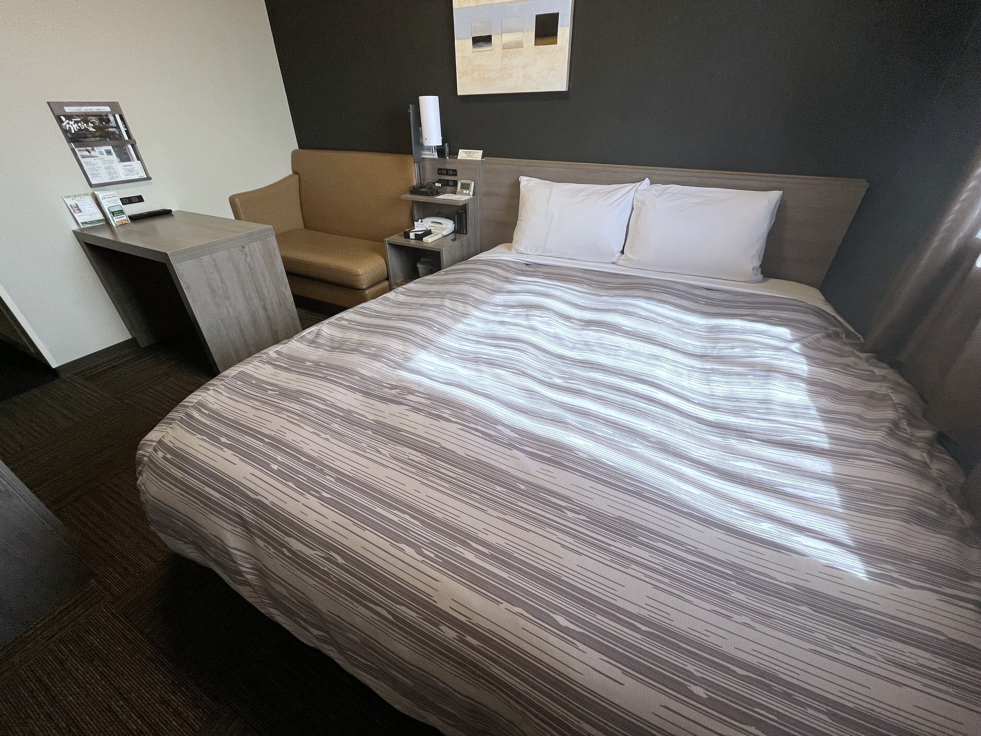 A hotel room with a large bed, a small desk, a chair, and a nightstand with a lamp.