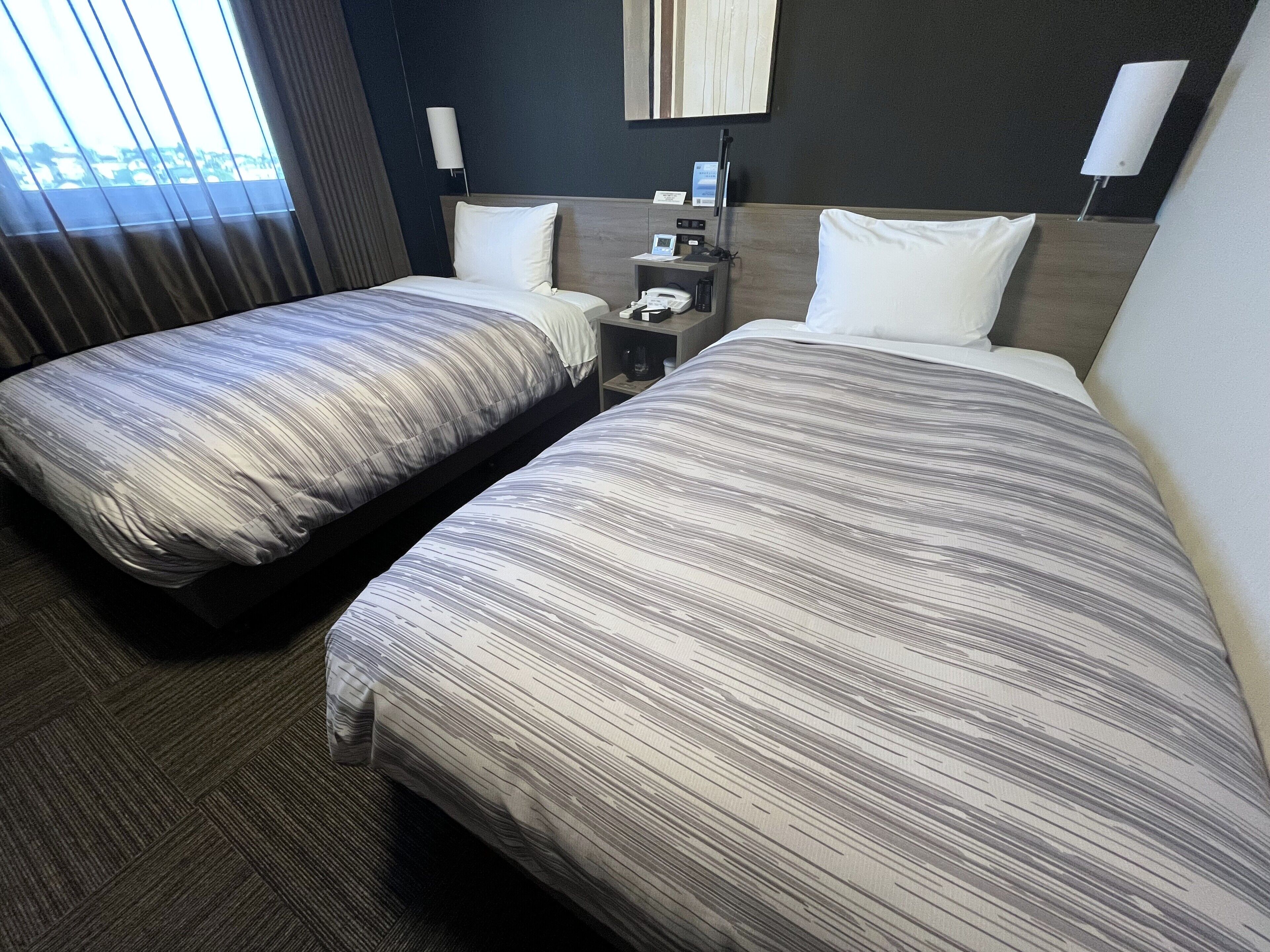 A hotel room with two beds, a nightstand, a lamp, and a window with curtains.