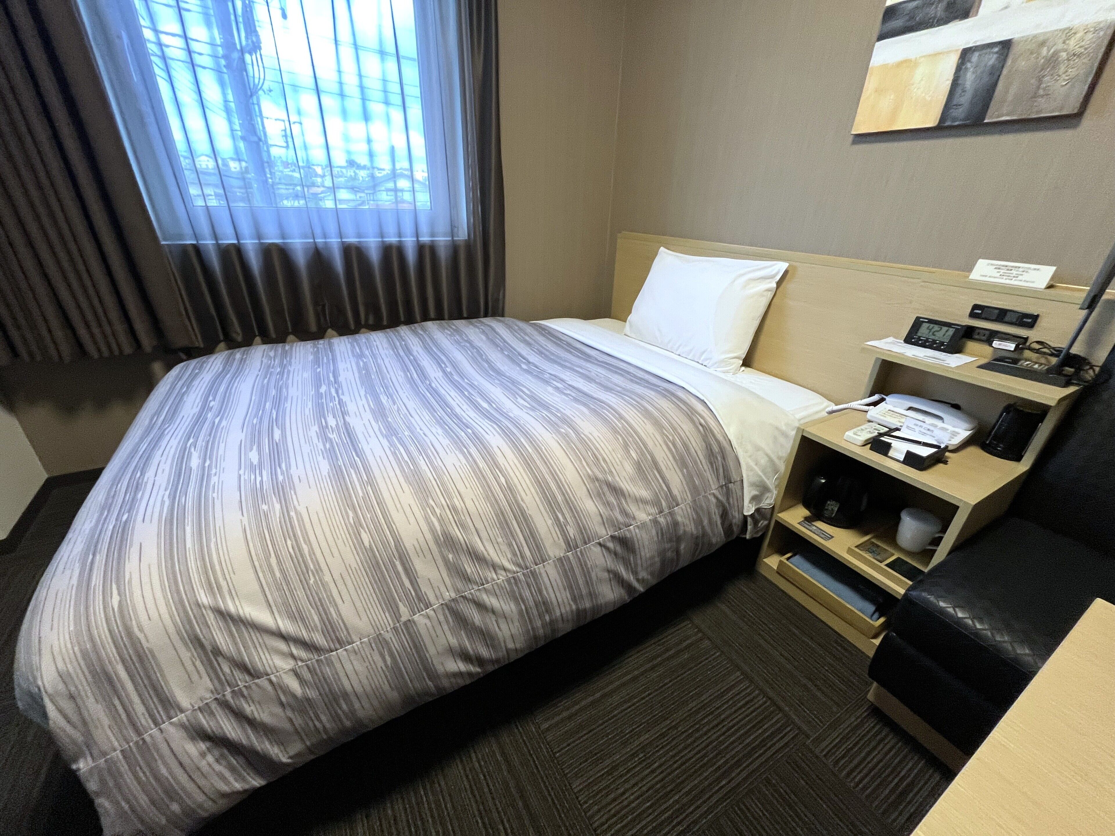 Habitación de hotel con cama, mesita de noche con teléfono y cafetera, ventana con persianas y una obra de arte colgada en la pared.