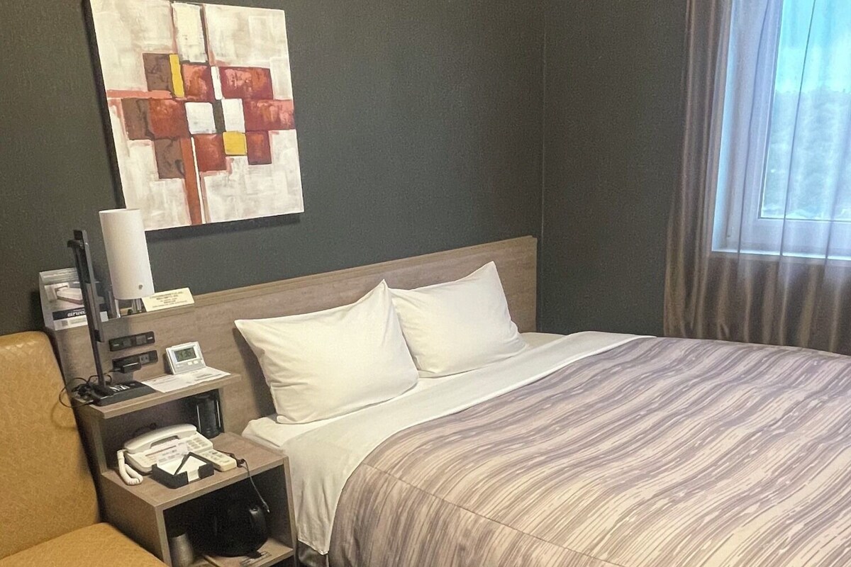A hotel room with a bed, a desk with a telephone, a lamp, and a wall-mounted artwork.