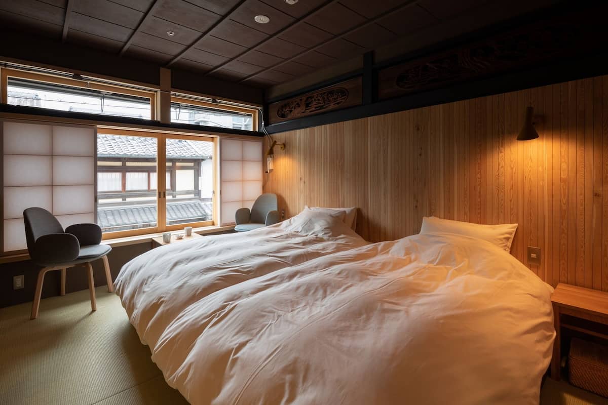 A traditional Japanese-style room with tatami flooring, a large bed with white bedding, a wooden headboard, a small table with a chair, and a window with shoji screens.