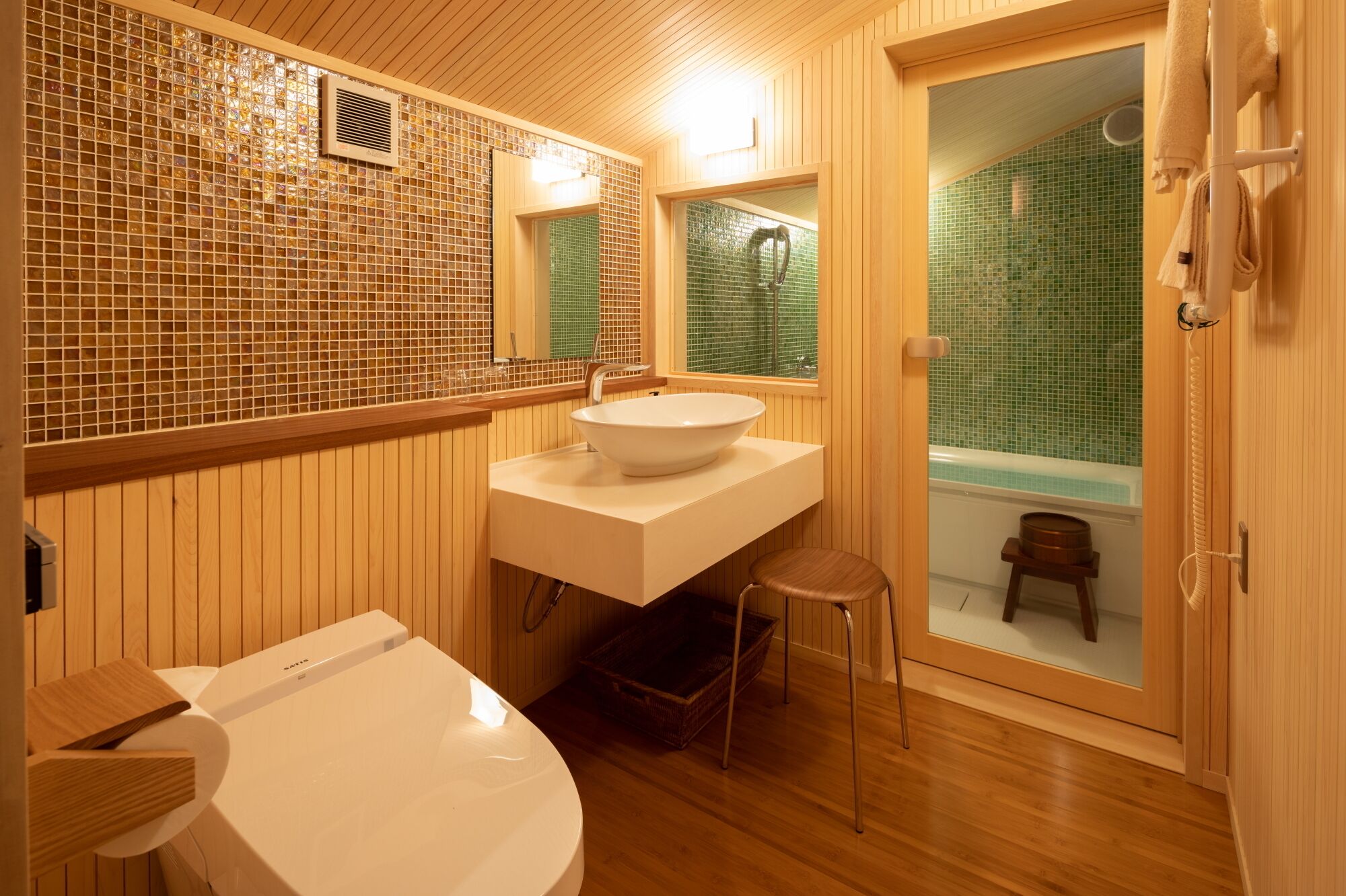 Un baño con tocador de madera, un lavamanos grande, un inodoro y una bañera visible a través de una puerta de cristal.