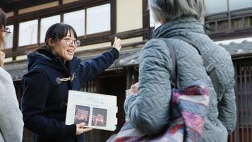 Eine Person hält ein Notizbuch mit Fotos und Text in der Hand und steht vor einem Gebäude mit traditioneller Holzbauweise.