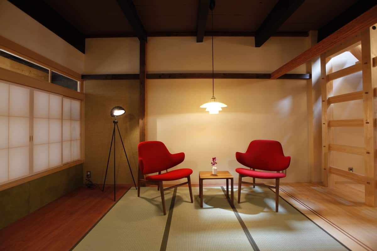 A traditional Japanese room with tatami flooring, two red chairs, a small wooden table, a floor lamp, and a sliding door.