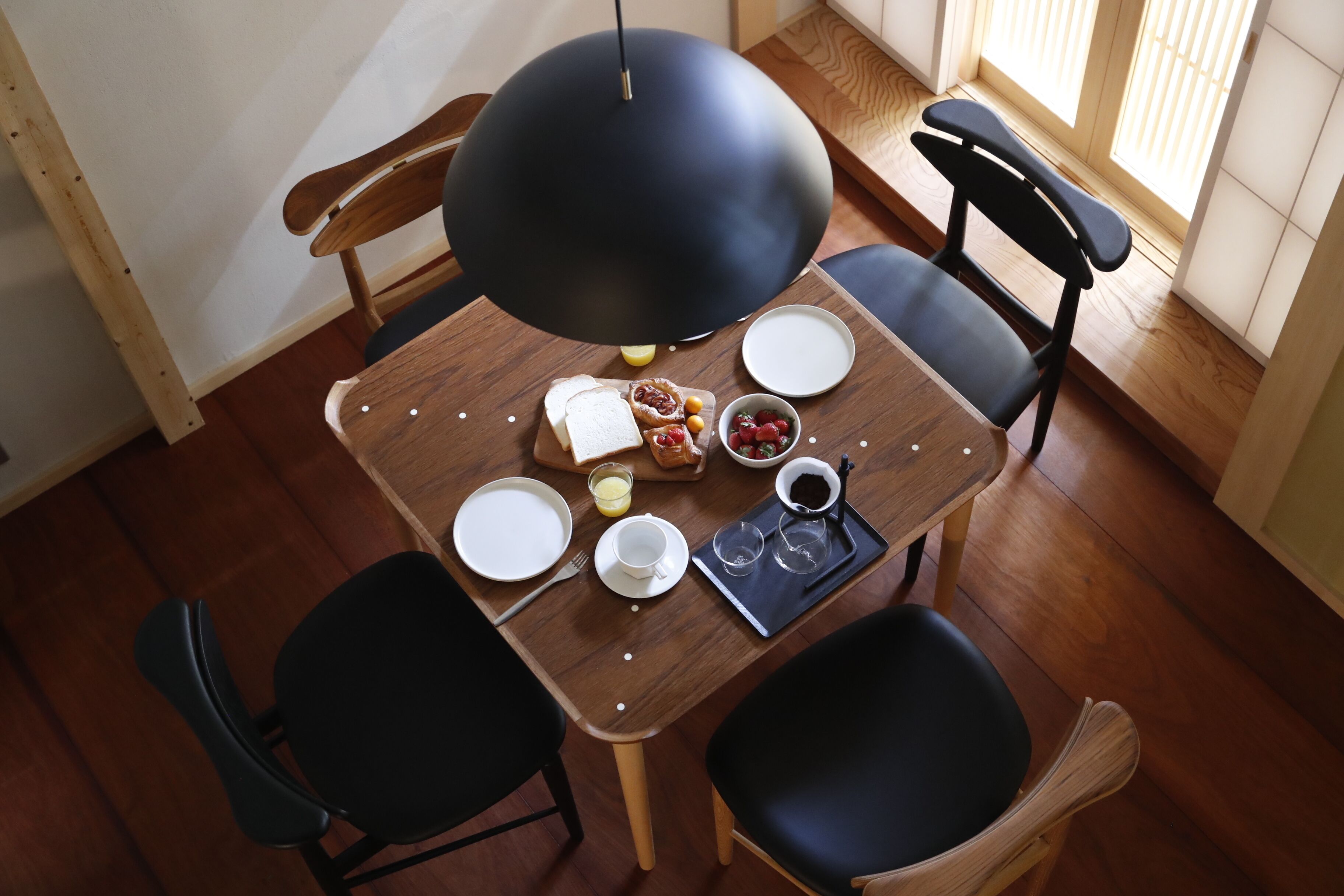 A dining table with a black pendant light, surrounded by four chairs, one of which is black. There are plates, cups, a bowl of fruit, and a tray with bread and a cup of coffee on the table.