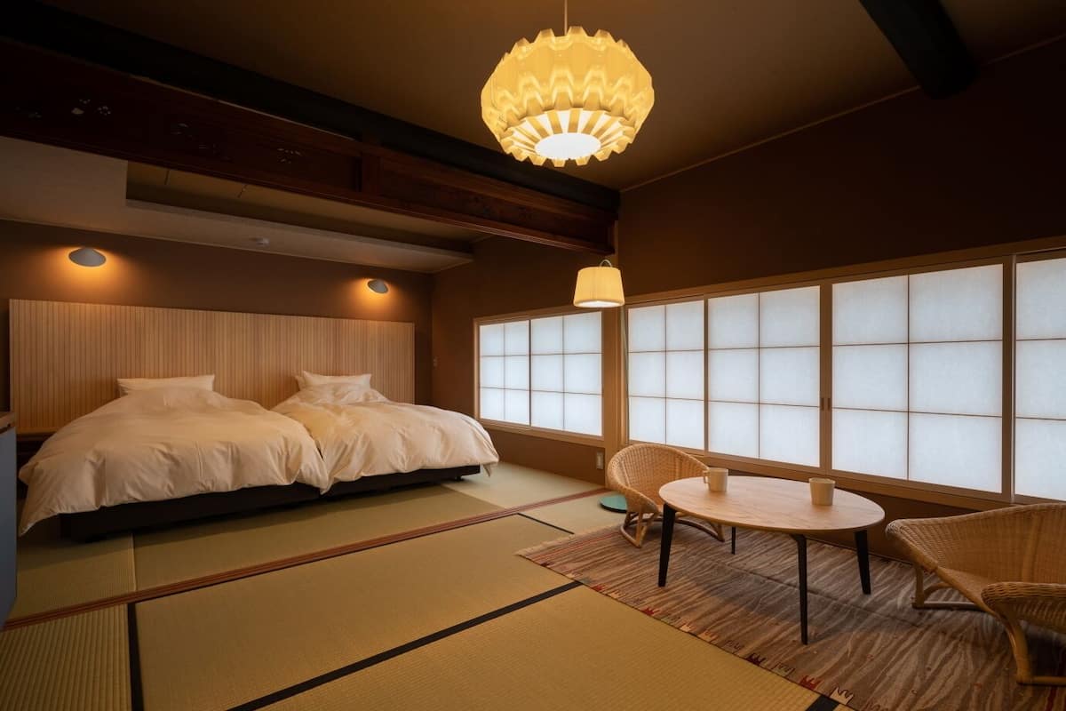 A traditional Japanese-style bedroom with tatami flooring, a low wooden bed, sliding shoji doors, and a round table with chairs.