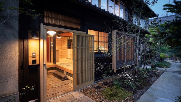 Una casa tradicional japonesa con fachada de madera, una entrada con puertas correderas y un pequeño jardín con rocas y plantas.