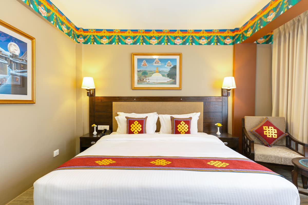 A hotel room with a large bed, two bedside lamps, a chair with a cushion, a framed picture on the wall, and a decorative ceiling.