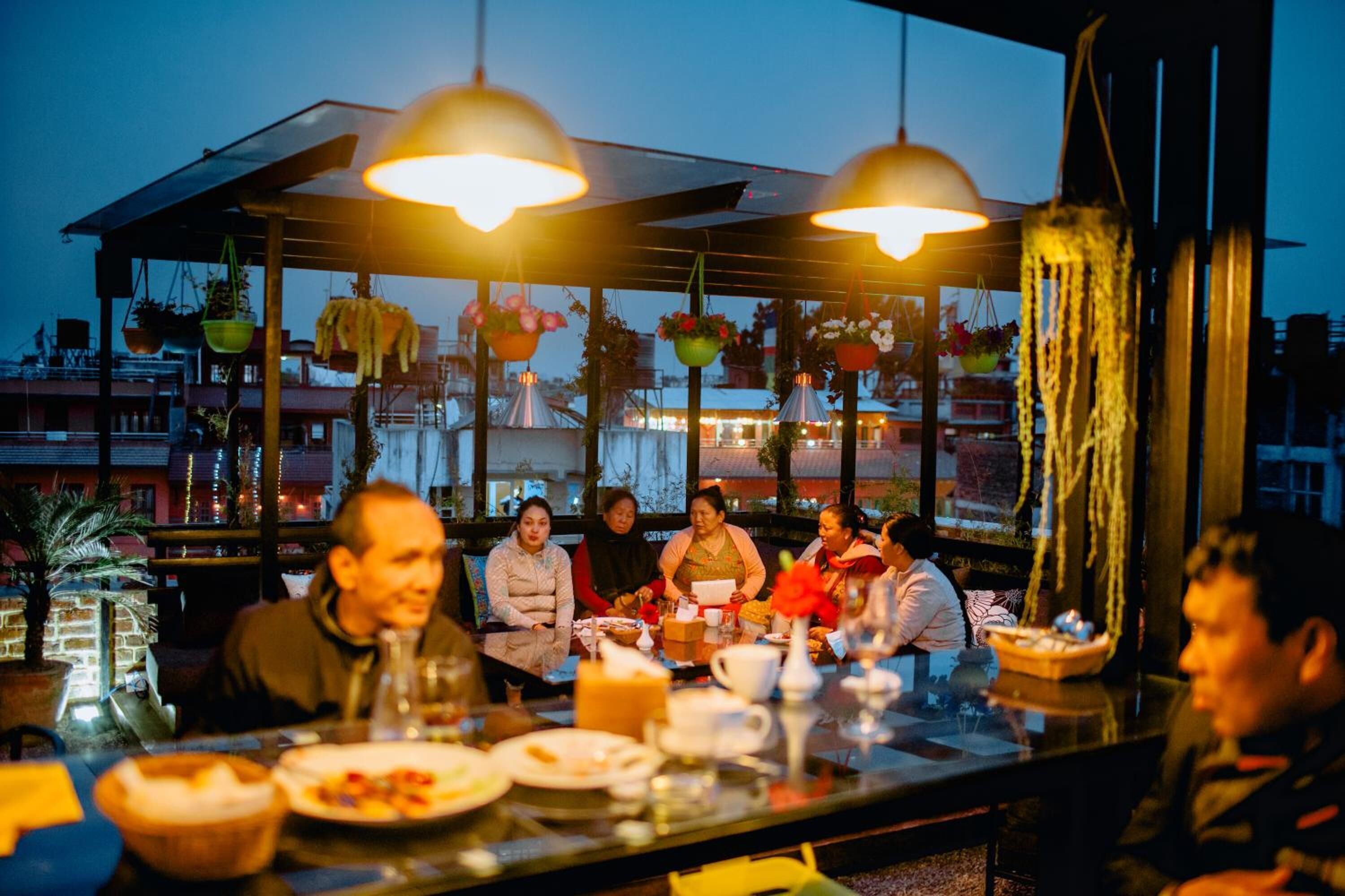 Un groupe de personnes est assis à une table dans un restaurant en plein air, décoré de plantes suspendues et d’éclairages suspendus.
