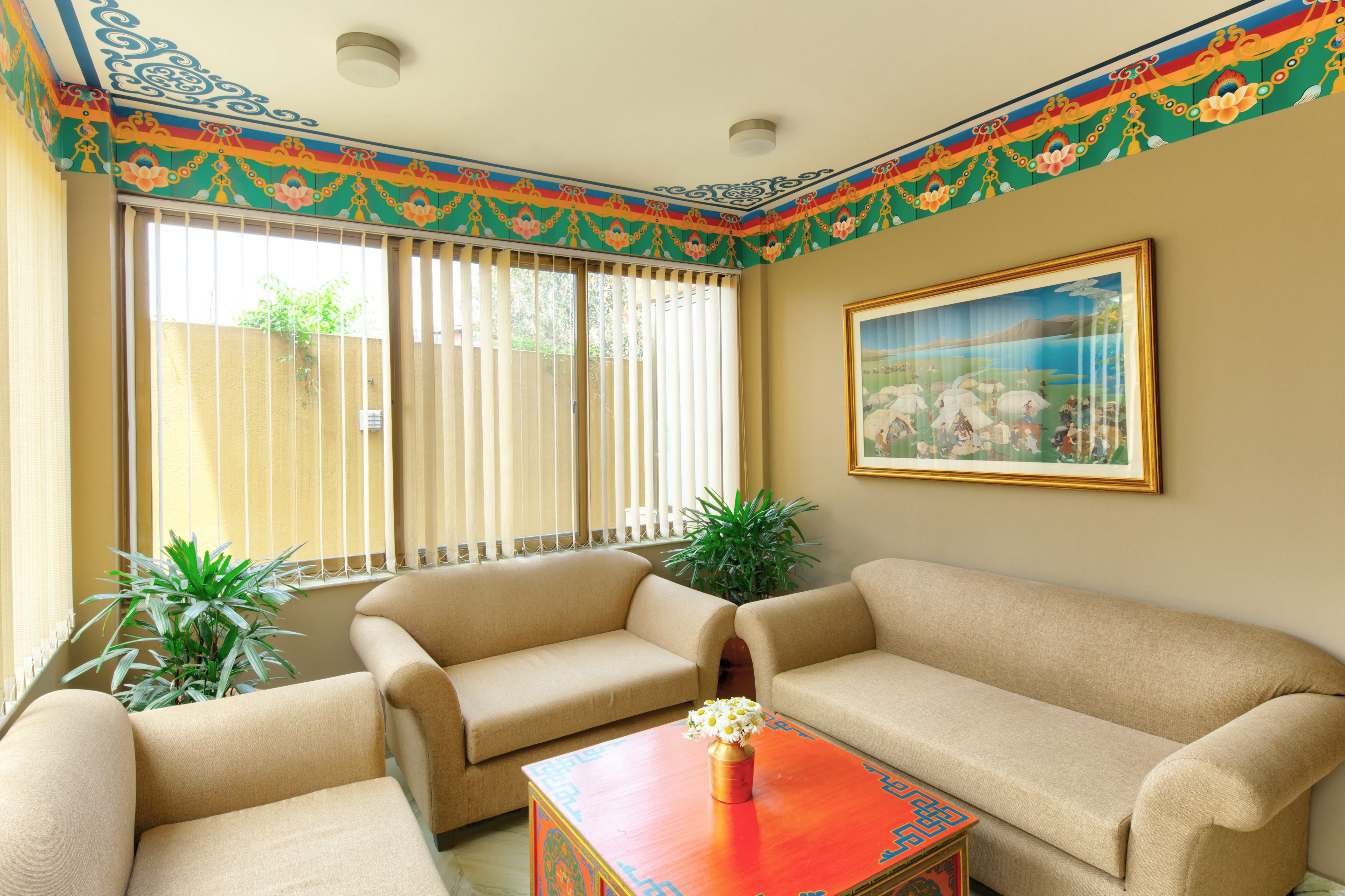A living room with beige sofas, a wooden coffee table with a vase, a painting on the wall, and a window with blinds.