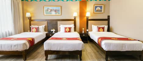 A hotel room with three single beds, each with a wooden headboard, white bedding, and a red decorative pillow. There are two bedside tables with lamps, a large window with curtains, and a patterned carpet.