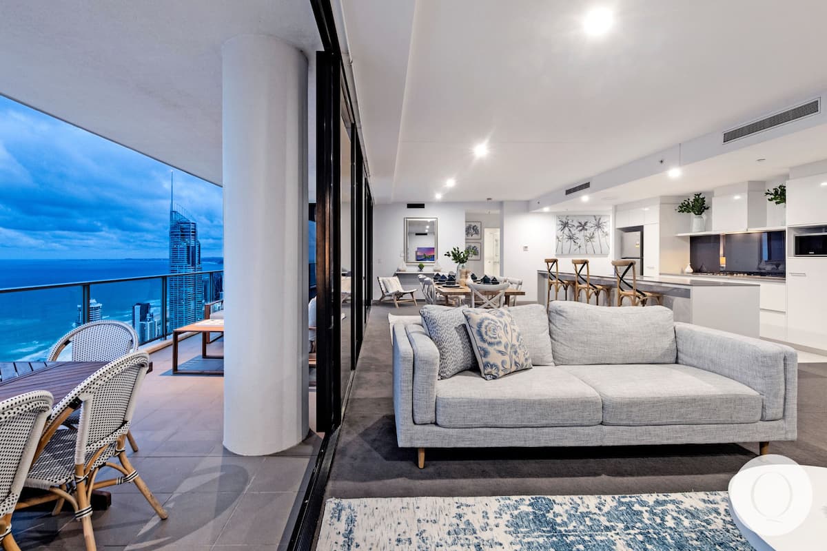 Uma sala de estar moderna com um sofá cinzento, uma área de jantar com cadeiras de madeira e uma cozinha com armários brancos.