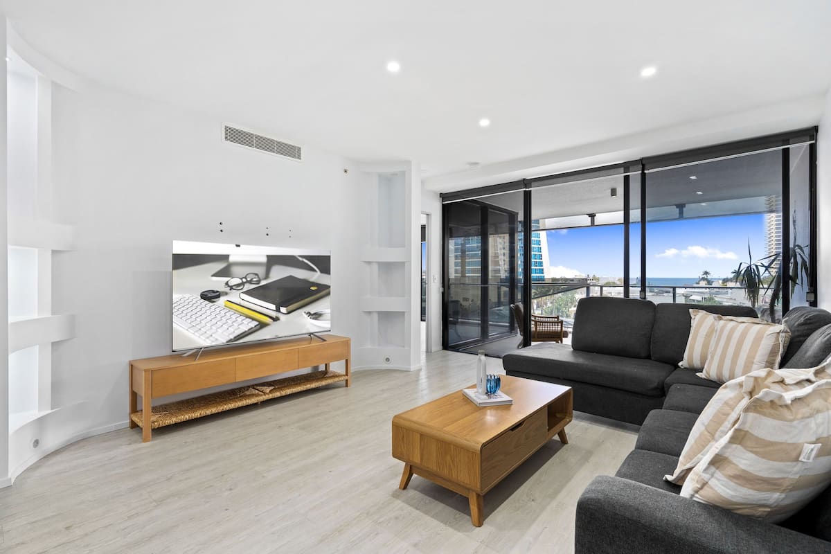 Uma sala de estar moderna com uma televisão de tela plana, uma mesa de centro de madeira, um sofá cinza e uma grande janela com vista para a cidade.