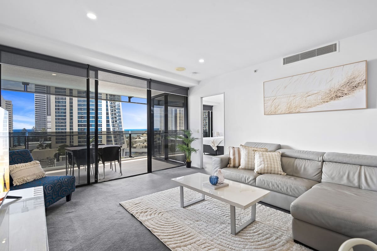 Uma sala de estar moderna com sofá, mesa de centro e vista para edifícios altos através de grandes portas de vidro.