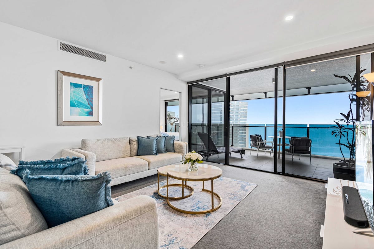 Sala de estar moderna com sofá, mesa de centro e vista para o mar através de grandes portas de vidro.
