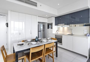 A modern kitchen with white cabinetry, stainless steel appliances, and a dining area with a white table and wooden chairs. - H'Residences - Q Stay (Surfers Paradise)