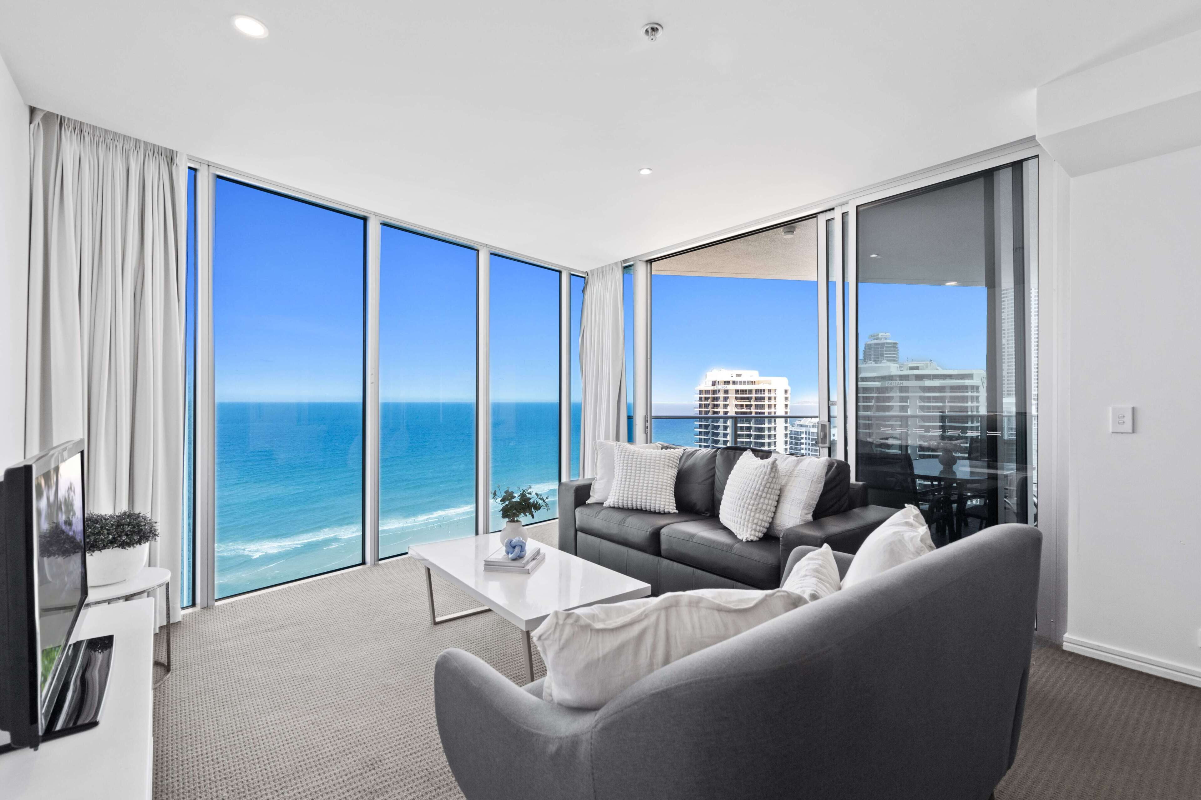 A modern living room with a large window overlooking the ocean, a gray sofa, a coffee table, and a flat-screen TV.
