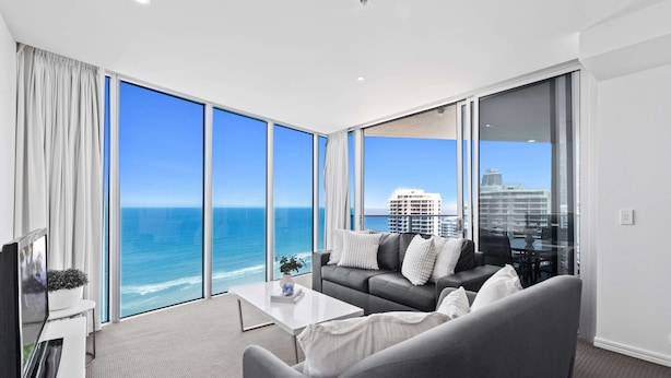 A modern living room with a large window overlooking the ocean, a gray sofa, a coffee table, and a flat-screen TV. - H'Residences - Q Stay (Surfers Paradise)