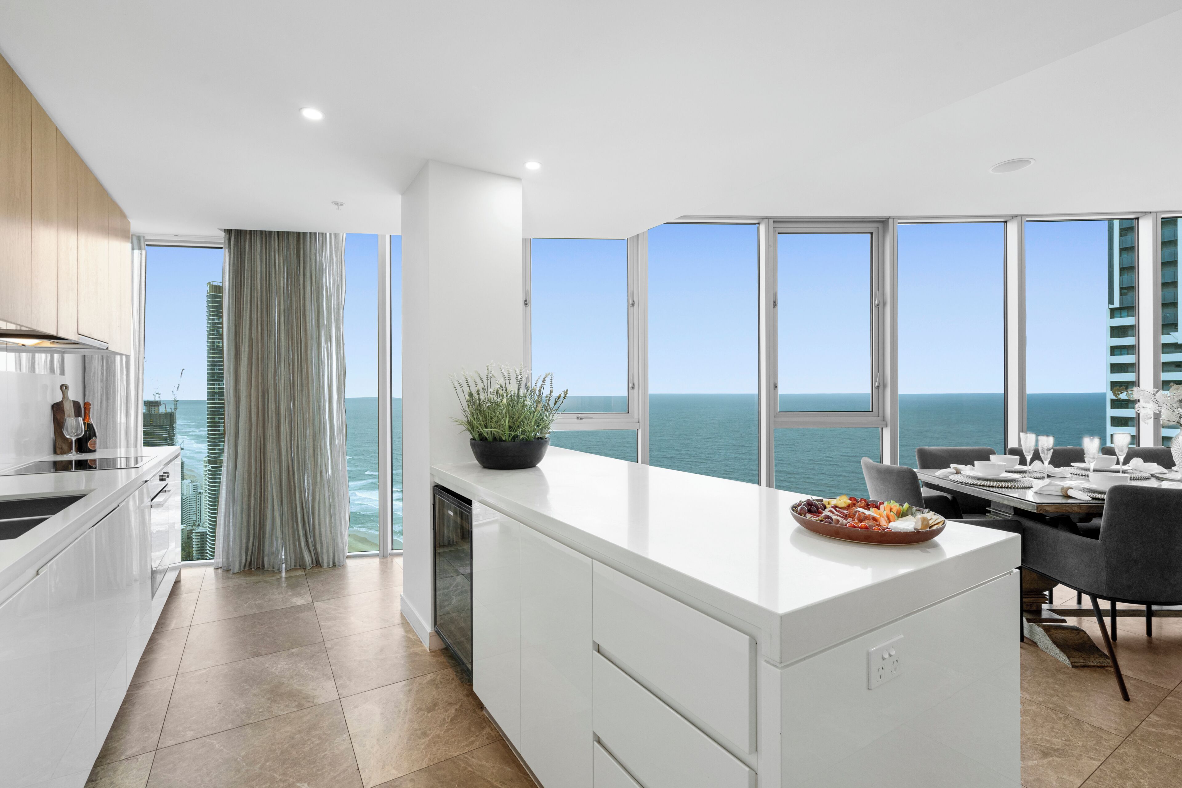 A modern kitchen with a large island, a dining area with a view of the ocean, and a balcony with a view of the city skyline.