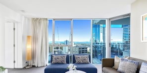 A modern living room with a sofa set, a coffee table, and a view of the cityscape through large windows. - H'Residences - Q Stay (Surfers Paradise)