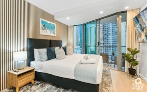 A modern bedroom with a large bed, a nightstand, a potted plant, and a view of high-rise buildings. - Legends - Q Stay (Surfers Paradise)