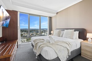 Une chambre d’hôtel avec un grand lit, un téléviseur fixé au mur, un banc en bois et une vue sur la ville grâce à de grandes fenêtres.
