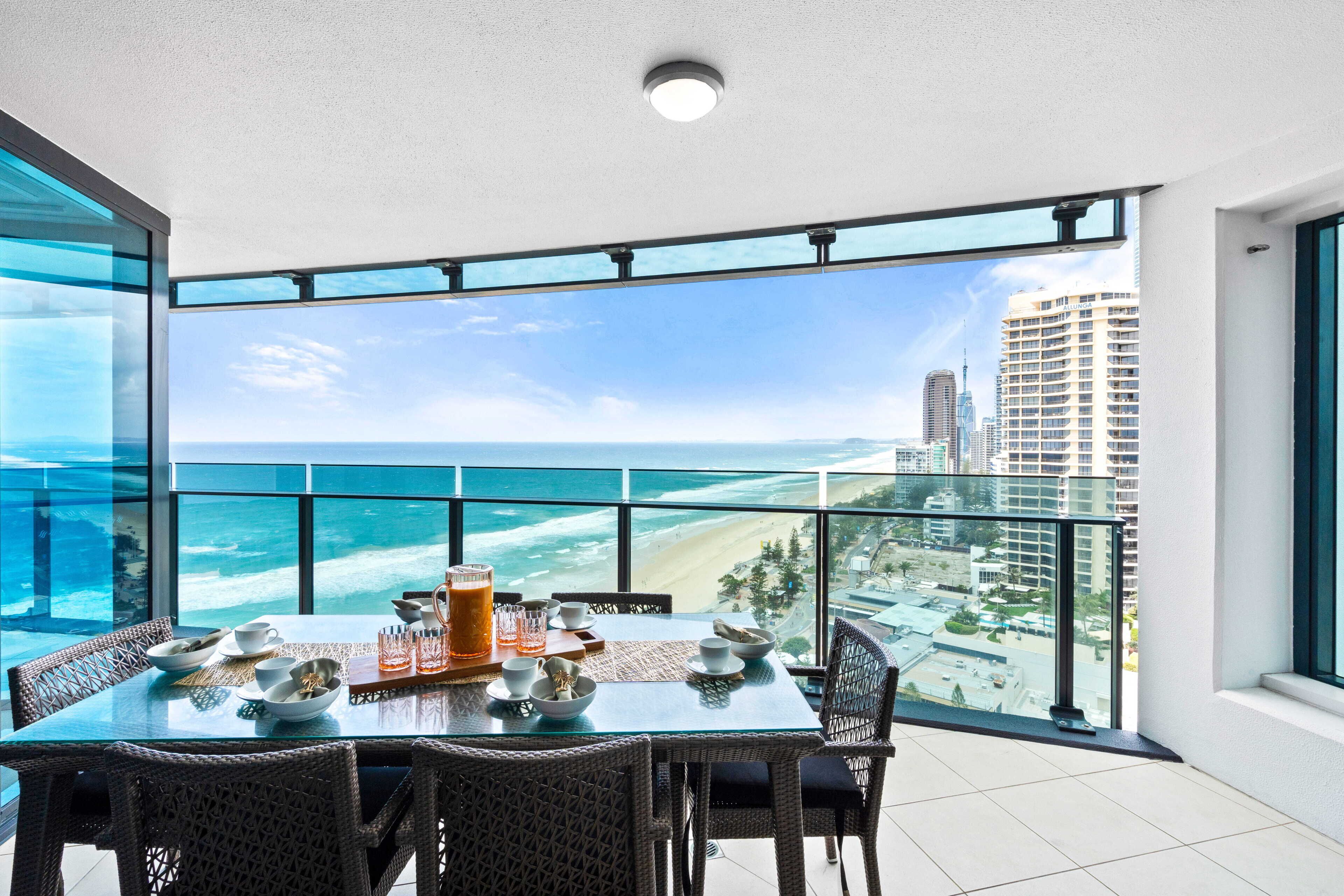 A dining area with a table set for four, overlooking a beach and cityscape through large windows.
