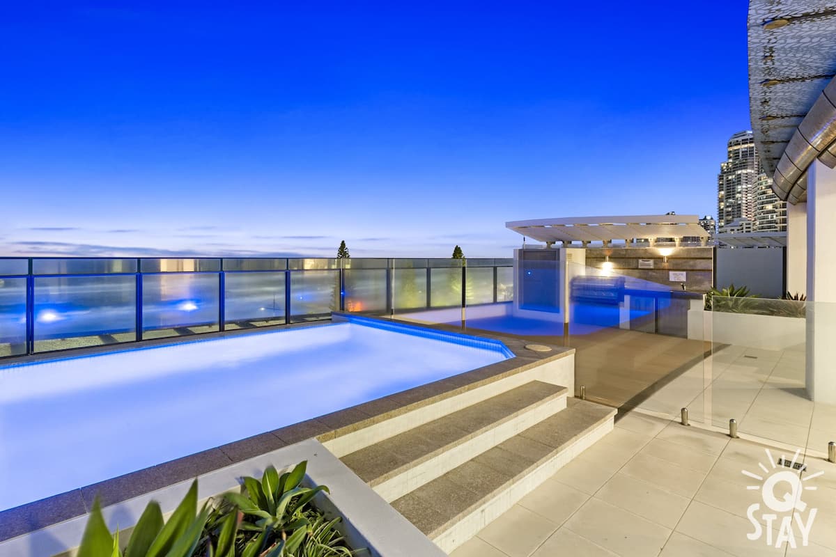 Ein Infinity-Pool auf dem Dach mit Glasgeländern, einer mit Steinplatten belegten Terrasse und Blick auf die Skyline der Stadt im Abendlicht.