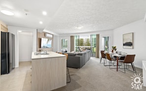 Un apartamento moderno con cocina, comedor y sala de estar. Cuenta con un ventanal grande, un televisor de pantalla plana y una mesa de comedor con sillas.