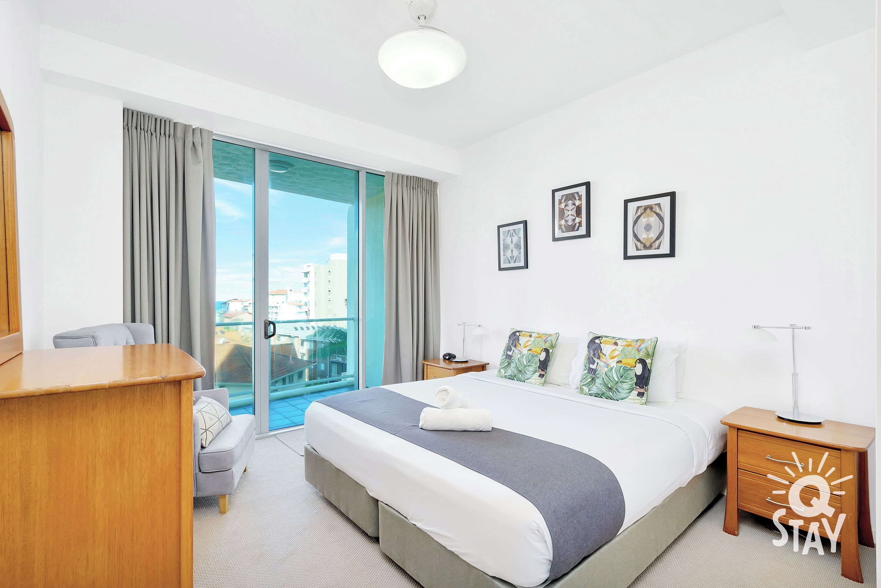 A hotel room with a large bed, a desk, a chair, a nightstand, and a view of the city through the balcony doors.