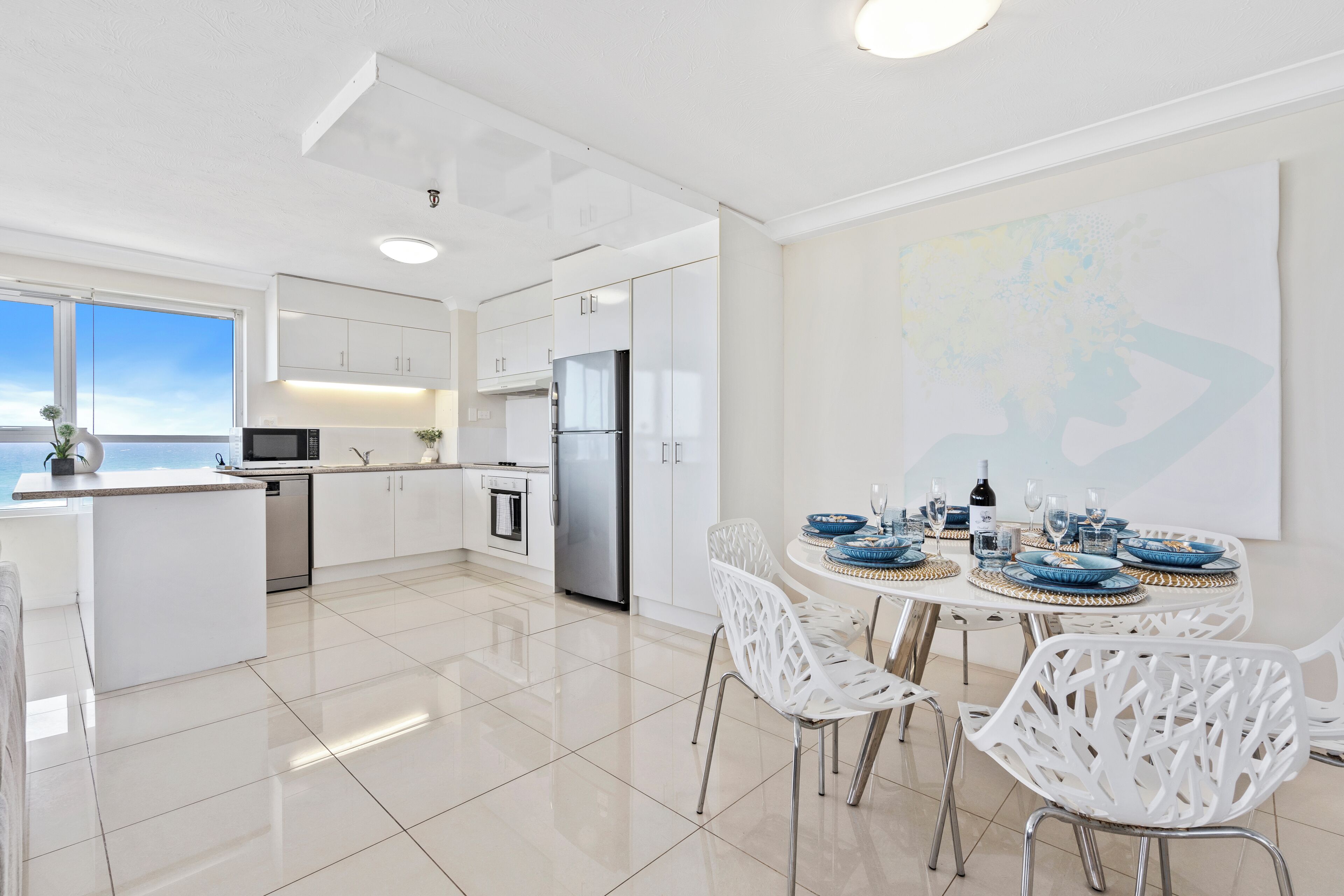A modern kitchen with white cabinetry, a dining area with white chairs, and a large window overlooking a body of water.