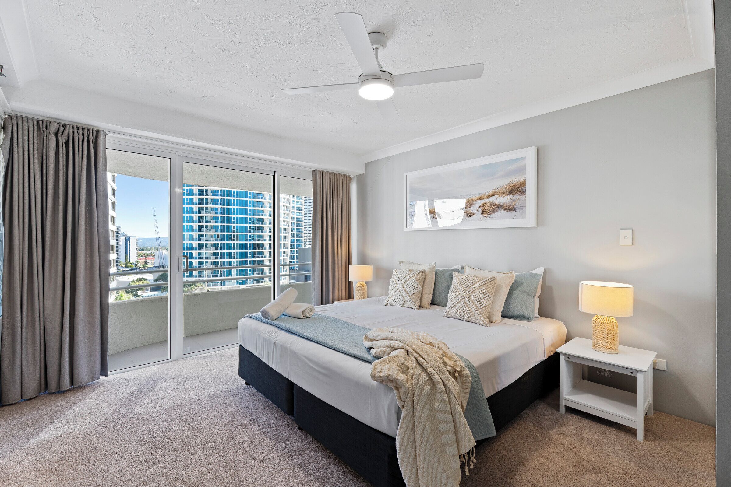 Une chambre à coucher comprenant un lit, des tables de chevet, un ventilateur de plafond, une fenêtre avec des rideaux et une vue sur la ville.