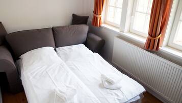 Un lit bien fait, avec du linge de lit et des serviettes blanches, un canapé gris et une fenêtre ornée de rideaux orange.