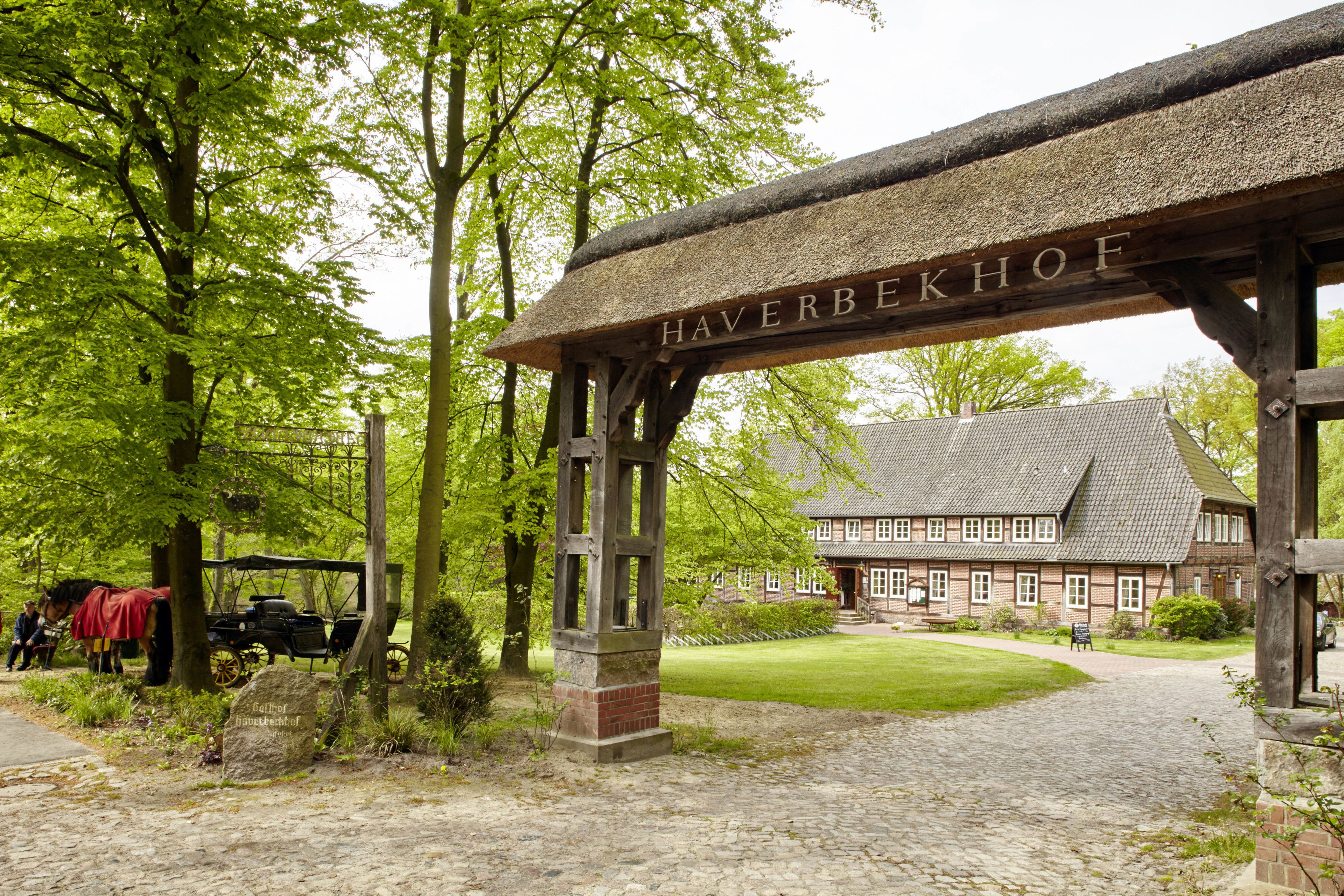 Ein traditioneller hölzerner Bogen mit der Aufschrift "Haverbekhof" darüber, der zu einem historischen Gebäude mit Ziegeldach und einem gepflegten Rasen führt.