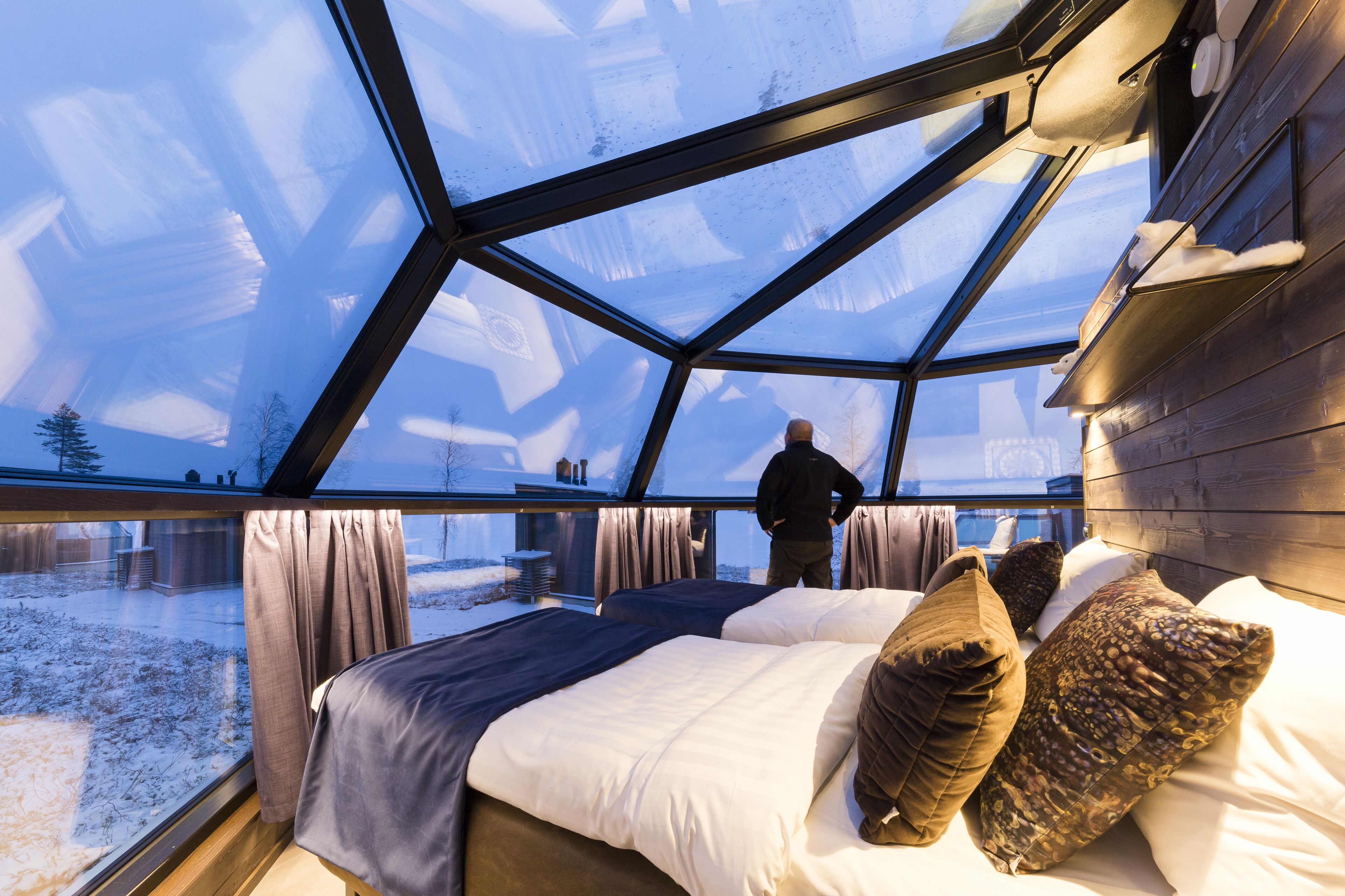 Un interior moderno con un amplio techo de vidrio, una cama con muchas almohadas, y una persona de pie junto a la ventana.
