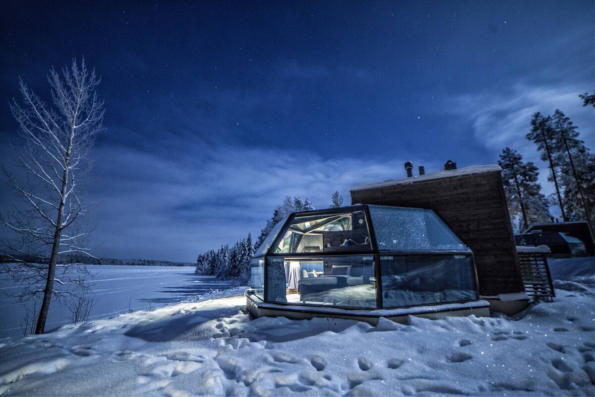 Une cabane en forme d’igloo, entièrement faite de verre et dont l’intérieur est visible, située dans un paysage enneigé sous un ciel nocturne dégagé.