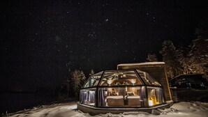 A snow-covered landscape with a glass-roofed cabin featuring a bed and a small table, surrounded by trees and a car.