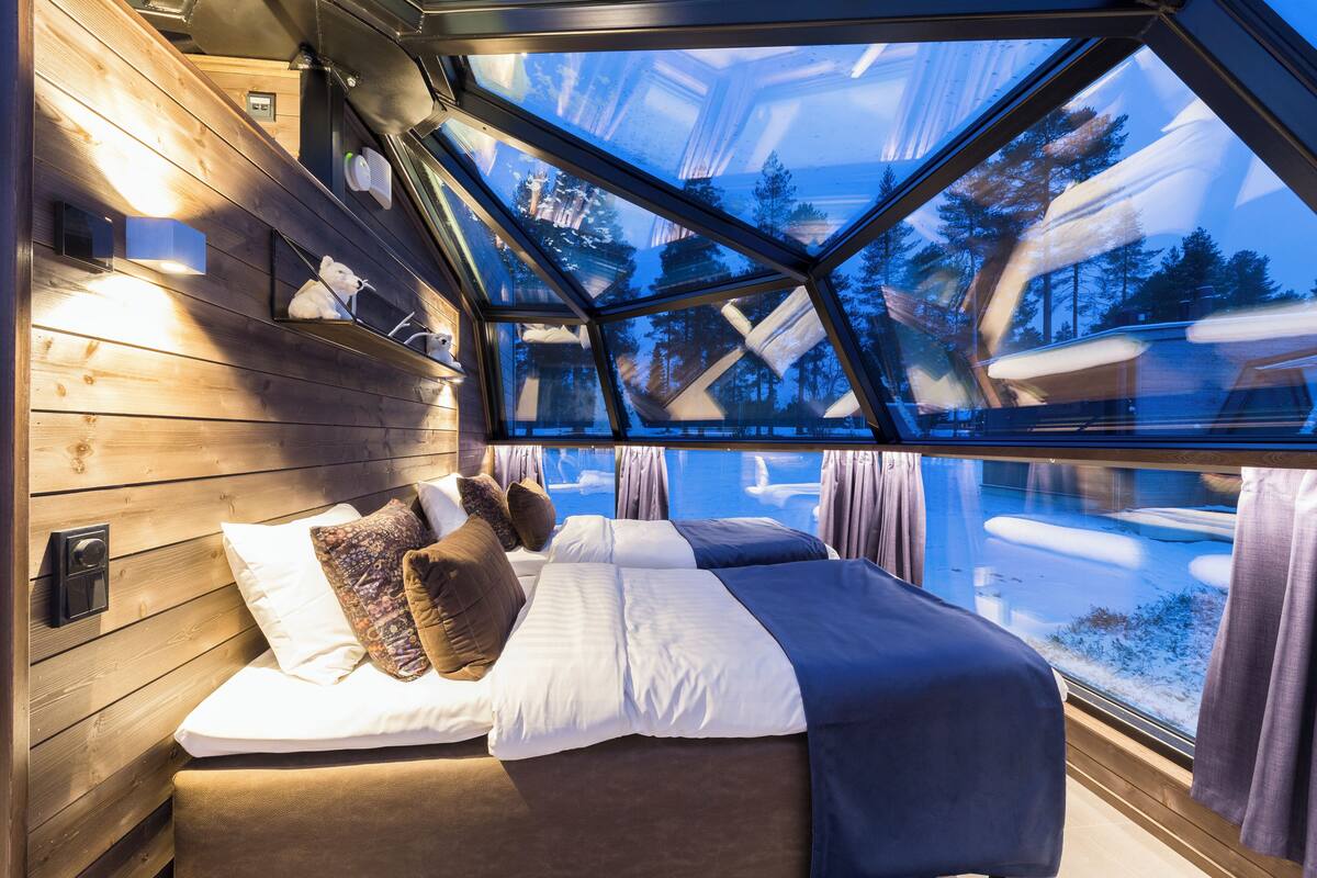 Une chambre avec une grande lucarne, des murs en bois, un lit recouvert de nombreux coussins et une vue sur un paysage enneigé.