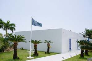 A modern building with a white exterior, a flagpole with a flag, and several palm trees in the foreground. - Windy Earth SILENT CLUB (Ishigaki)