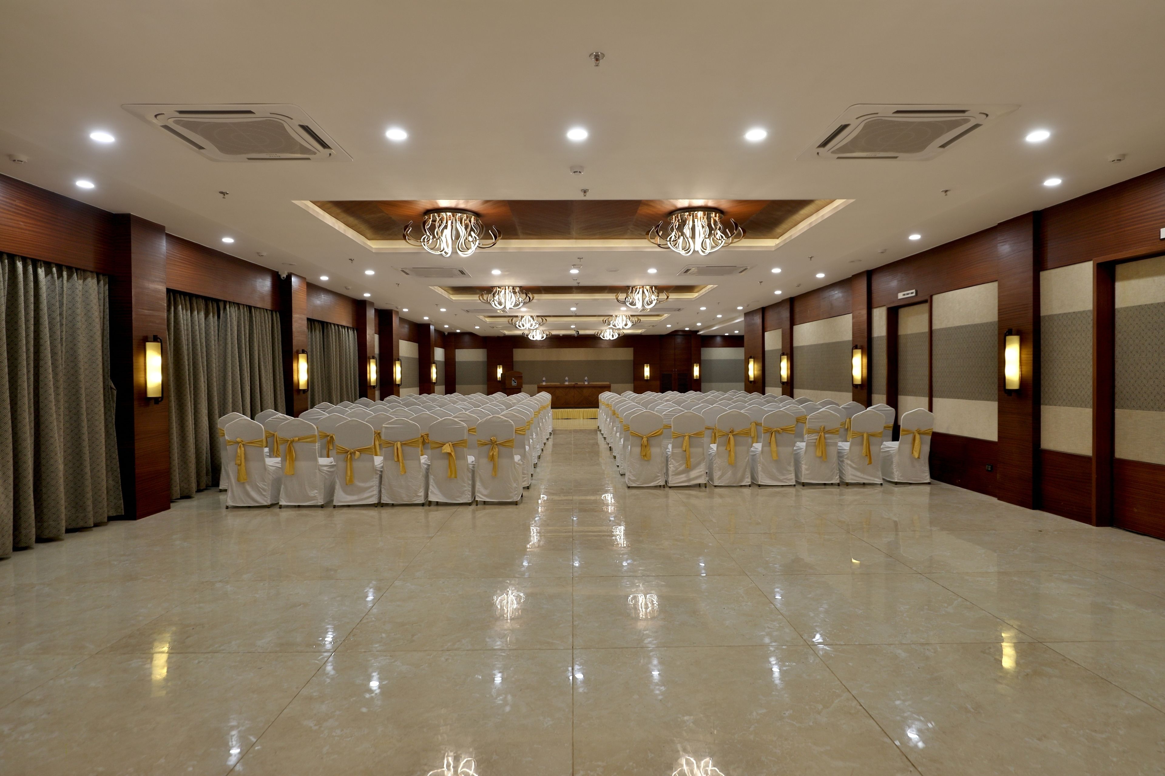 A spacious conference room with rows of white chairs, chandeliers, and wall sconces.