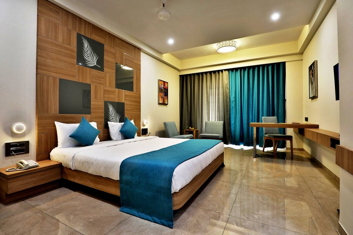A modern hotel room with a large bed, a desk, and a chair. There is a small table with a lamp, a telephone, and a wall-mounted light.