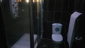 A small, dark bathroom with a toilet, a shower enclosure, and a towel hanging on the wall.