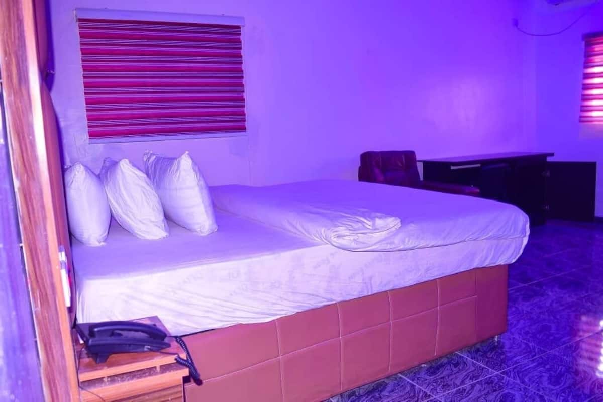 Een slaapkamer met een bed, kussens, een nachtkastje en een raam met jaloezieën.