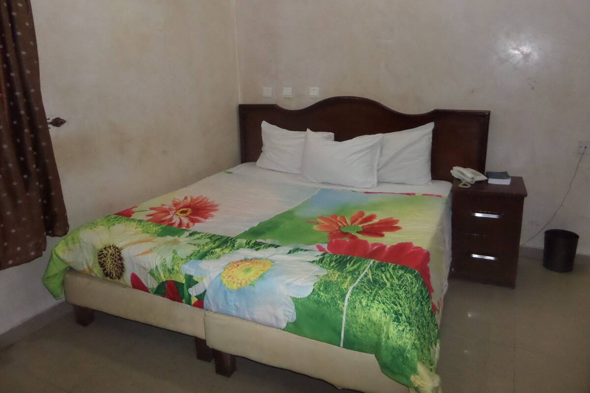A bed with a floral bedspread, a wooden headboard, white pillows, a bedside table with a phone, and a trash bin.