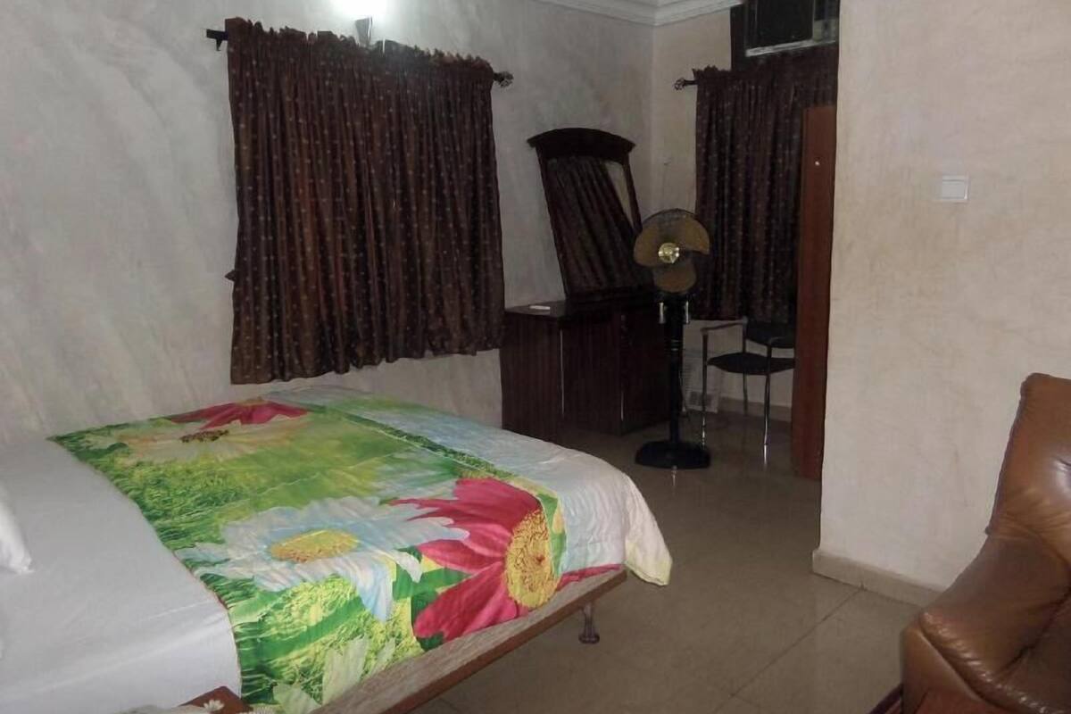 A bedroom with a bed, a chair, a desk, and a fan.