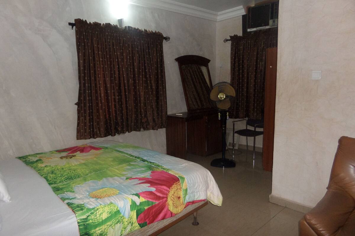 A bedroom with a bed, a desk, a chair, a mirror, and a fan.