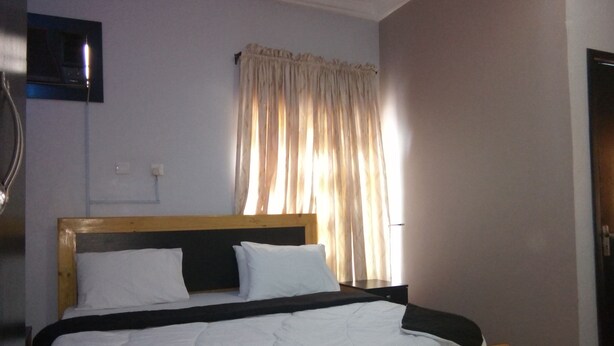 Una cama individual con ropa de cama blanca, cabecera de madera y ventana con cortinas translúcidas.